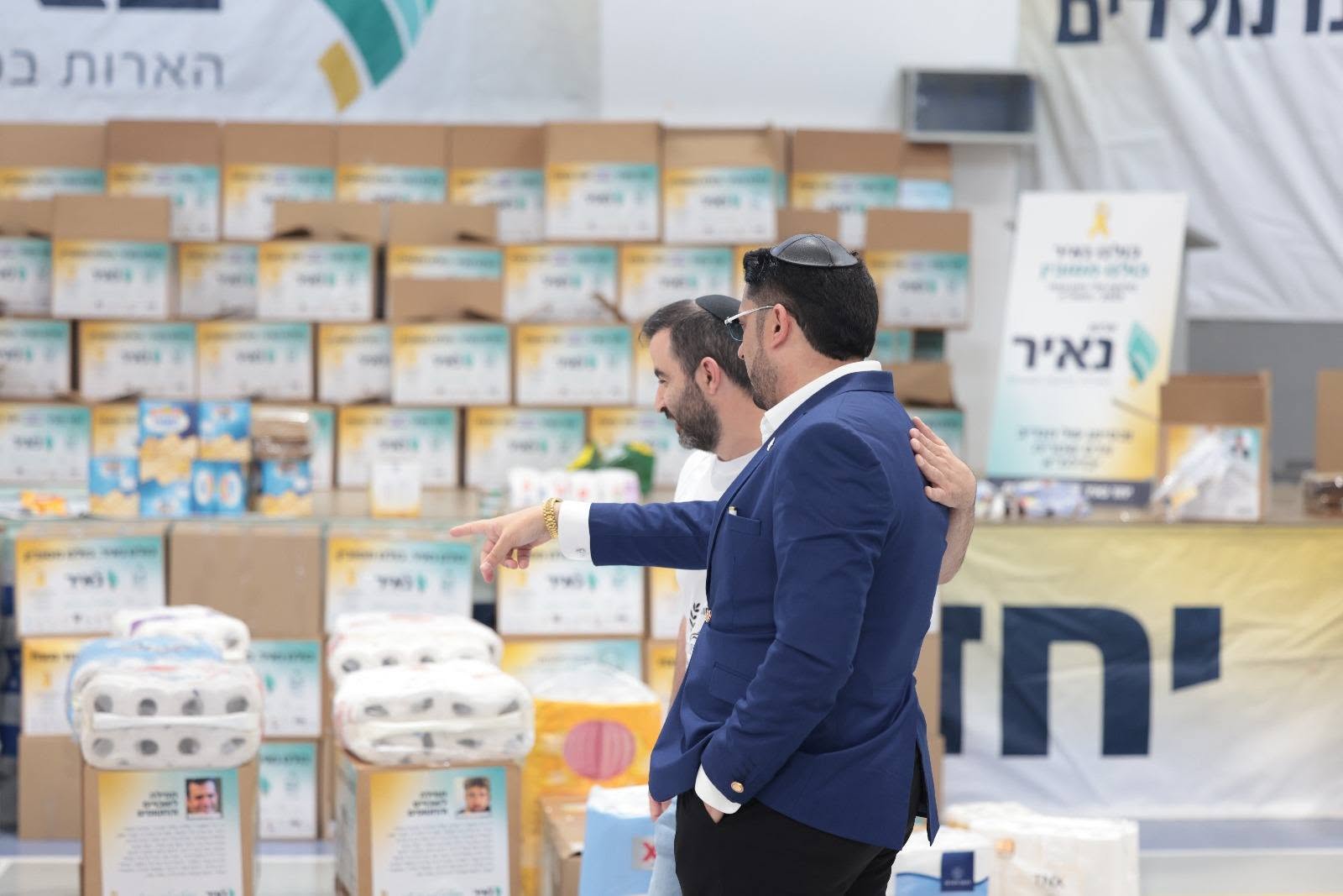 אדם שטרית ומשתתף נוסף עומדים מול סלי מזון ארוזים באולם